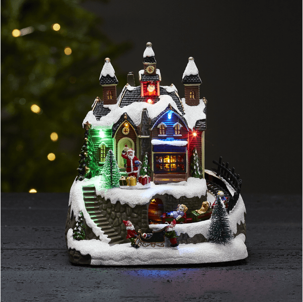 Trainville Mehrfarbig Von Star Trading - Dekoratives Weihnachtsdorf bestehend aus Häusern mit Türmen, hellen und lauten Fenstern und einem stehenden und winkenden Weihnachtsmann.